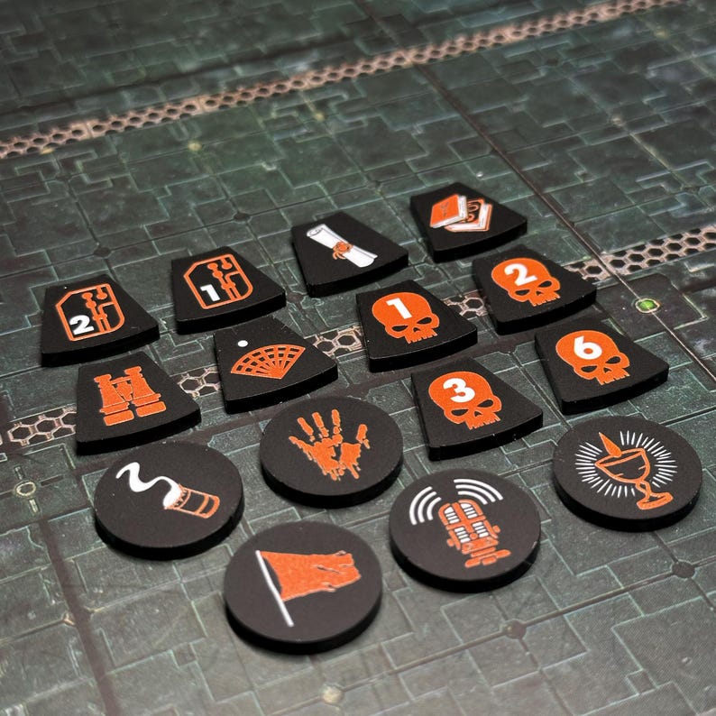 Puede incluir: Una colecci&oacute;n de fichas de juego negras con ilustraciones y n&uacute;meros naranjas. Las fichas presentan varios s&iacute;mbolos, incluyendo calaveras, una huella de mano, una bandera y un micr&oacute;fono. Las fichas est&aacute;n dispuestas sobre una superficie texturizada de color verde oscuro.