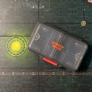 Puede incluir: Un contenedor de pl&aacute;stico transparente con las palabras "BROKEN TOKEN" en letras naranjas. El contenedor es rectangular con m&uacute;ltiples compartimentos y un asa roja. El contenedor est&aacute; sobre una superficie verde y gris.