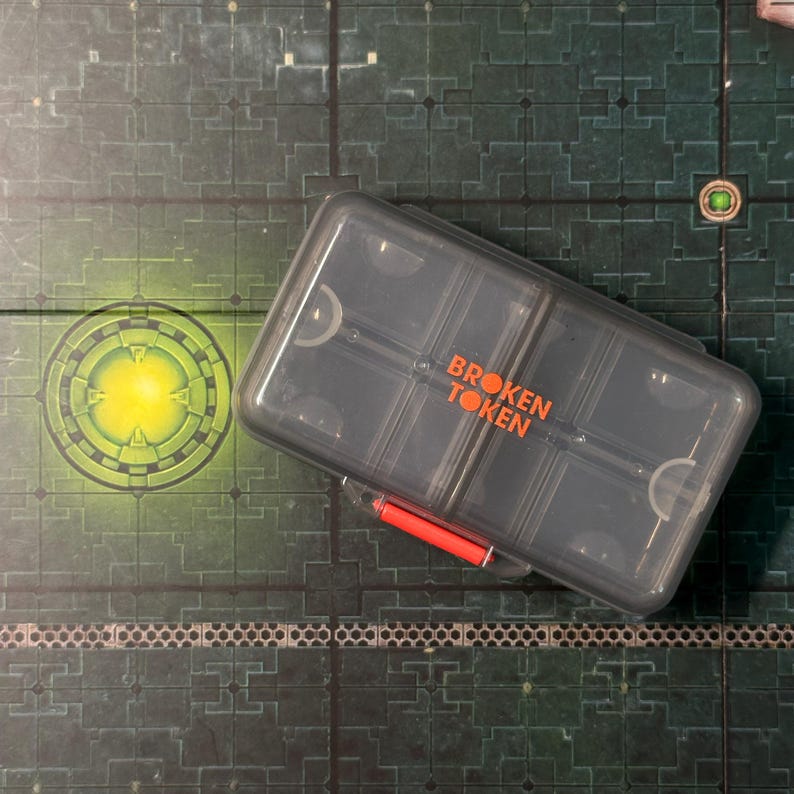 Puede incluir: Un contenedor de pl&aacute;stico transparente con las palabras "BROKEN TOKEN" en letras naranjas. El contenedor es rectangular con m&uacute;ltiples compartimentos y un asa roja. El contenedor est&aacute; sobre una superficie verde y gris.