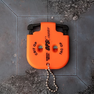 May include: Bright orange OPS Counter by Broken Token. The counter has two black handles and a small metal chain. The text "GRIT OP" and "TAC OP" are printed on the sides. The text "OPS Counter" and "BROKEN TOKEN" are printed on the front.