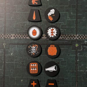 Puede incluir: Una colecci&oacute;n de fichas de juego negras con gr&aacute;ficos naranjas y blancos. Las fichas presentan varios s&iacute;mbolos, incluyendo una pistola, una gota, una bomba y un meg&aacute;fono. Algunas fichas tienen las letras "AP" y un s&iacute;mbolo m&aacute;s o menos.