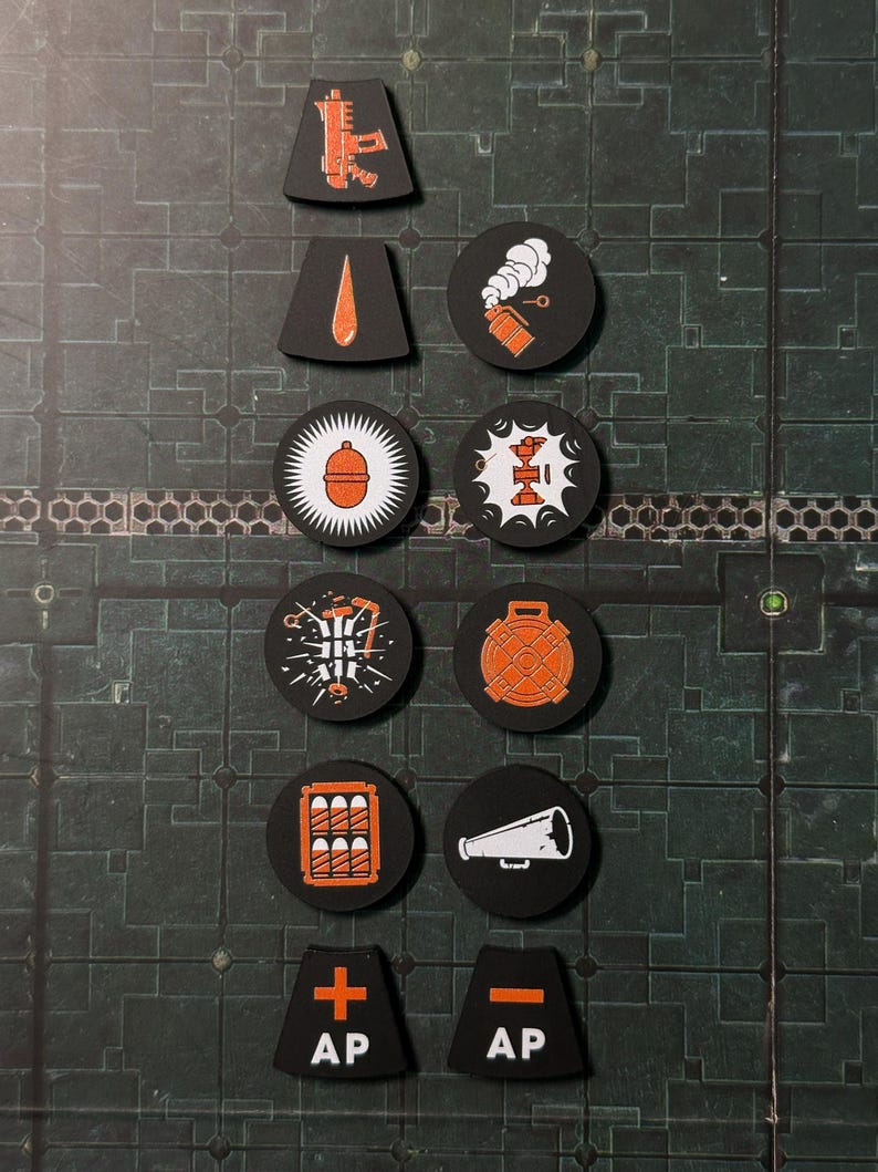 Puede incluir: Una colecci&oacute;n de fichas de juego negras con gr&aacute;ficos naranjas y blancos. Las fichas presentan varios s&iacute;mbolos, incluyendo una pistola, una gota, una bomba y un meg&aacute;fono. Algunas fichas tienen las letras "AP" y un s&iacute;mbolo m&aacute;s o menos.