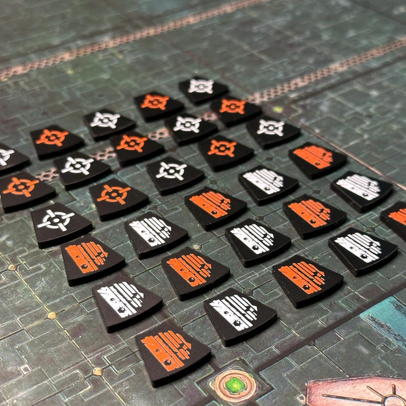 Puede incluir: Una colecci&oacute;n de fichas de juego negras en forma de escudo con varios s&iacute;mbolos blancos y naranjas. Las fichas est&aacute;n dispuestas sobre una superficie texturizada, verde y gris, lo que sugiere un juego de mesa. Los s&iacute;mbolos incluyen miras y formas geom&eacute;tricas.
