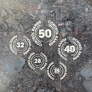 May include: A set of clear, circular game tokens with the words "Control Range" printed around the edges. Each token displays a different number: 25, 28, 32, 40, and 50. The tokens are on a textured, dark surface.