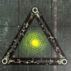 Puede incluir: Una pieza de juego triangular negra con diseños de rayos y acentos circulares blancos. La pieza está colocada sobre un tablero de juego verde y gris con un centro amarillo brillante.