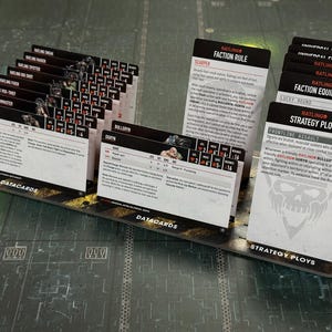 Könnte beinhalten: Eine Sammlung von schwarz-weißen Datacards für ein Tabletop-Spiel, mit Text und Illustrationen. Die Karten sind in einer gestaffelten Anordnung angeordnet, wobei einige Karten mit "Fraktionsregel" und "Strategie-Trick" beschriftet sind. Die Karten liegen auf einer strukturierten Oberfläche.