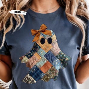 Pode incluir: Camiseta azul denim com um design de fantasma de patchwork e um laço marrom. O fantasma é feito de vários tecidos estampados em tons de azul, laranja e verde. A palavra "DENIM" está impressa na camiseta.