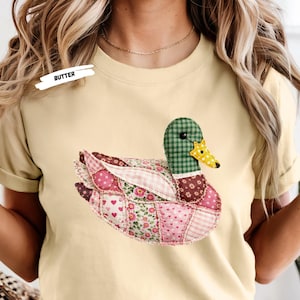 以下が含まれることがあります： バター色のTシャツ。パッチワークのアヒルのデザイン。アヒルは緑のチェック柄の頭、黄色と黒のくちばし、ピンクと花柄の生地でできた体を持っています。「BUTTER」の文字が白いラベルに印刷されています。