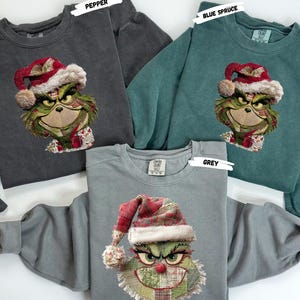 May include: Three crewneck sweatshirts in pepper, blue spruce, and grey. Each sweatshirt features an embroidered Grinch face wearing a Santa hat. The Grinch's face is green, with a red nose and a red and white hat.