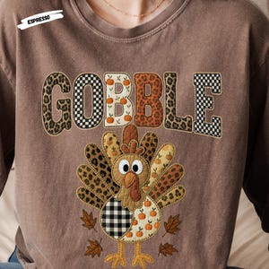 以下が含まれることがあります： 「GOBBLE」の文字がパッチワークデザインで施された茶色の長袖シャツ。「O」の文字はカボチャで装飾されています。「GOBBLE」の下には、パッチワークデザインの七面鳥のグラフィックがあります。シャツには「ESPRESSO」と書かれています。
