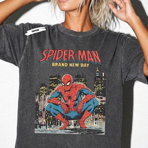 May include: A charcoal gray short-sleeve t-shirt featuring a graphic print of Spider-Man crouching in front of a city skyline. The text reads "SPIDER-MAN" in red and "BRAND NEW DAY" in yellow, with a small "PEPPER" label near the shoulder.
