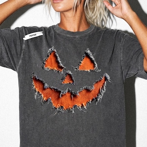 Puede incluir: Una camiseta de manga corta gris carbón con un gráfico de cara de calabaza desgastada con bordes deshilachados que revelan una tela naranja brillante debajo. La palabra PEPPER está impresa en letras blancas pequeñas cerca del hombro izquierdo.