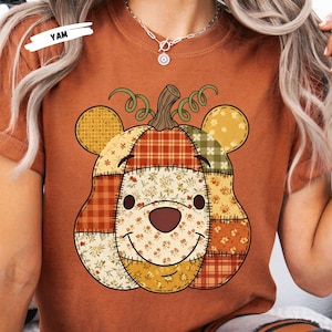 May include: Rust-colored t-shirt featuring a patchwork pumpkin design with bear ears and a smiling face. The pumpkin is made of various patterned fabrics in fall colors. The word "YAM" is visible in the upper left corner.
