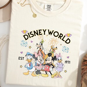 Puede incluir: Camiseta de color crema con las palabras "DISNEY WORLD" sobre un gr&aacute;fico de personajes de Disney, incluyendo Mickey Mouse, Minnie Mouse, Donald Duck, Goofy, Pluto y Daisy Duck. El texto "EST 1971" tambi&eacute;n est&aacute; impreso en la camiseta.
