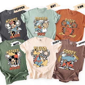 May include: Six different colored t-shirts featuring Disney characters. Each shirt has a different character graphic, including Minnie Mouse, Donald Duck, Daisy Duck, Pluto, Mickey Mouse, and Goofy. The shirts are in various colors, such as gray, green, and brown.