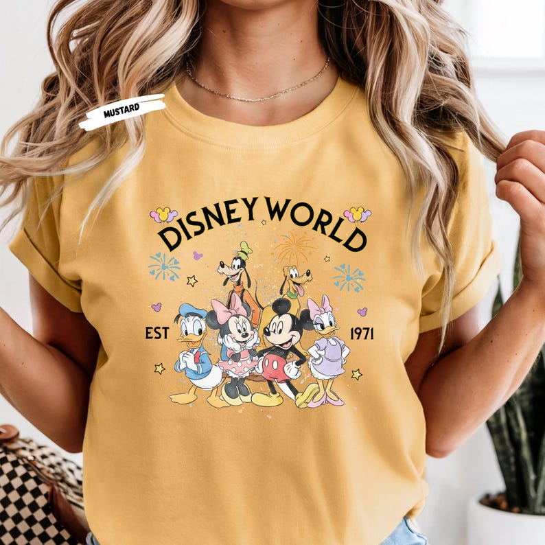 Puede incluir: Camiseta de color mostaza con un gr&aacute;fico de Disney World con Mickey Mouse, Minnie Mouse, Donald Duck, Daisy Duck, Goofy y Pluto. El gr&aacute;fico incluye el texto "Disney World" y "EST 1971".