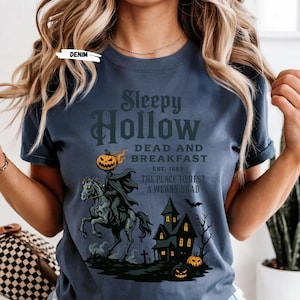 "Sleepy Hollow Halloween shirt featuring a vintage-style Headless Horseman on a spooky horse holding a jack-o'-lantern in front of a haunted house with glowing pumpkins and bats. This eerie Halloween graphic tee is printed on a moss green t-shirt and perfect for spooky season, trick-or-treat, horror movie lovers, gothic fall fashion, haunted house fans, or those searching for a unique October outfit. Ideal for fall events, pumpkin patches, or Halloween parties."
