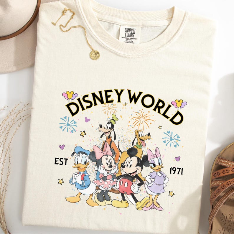 Puede incluir: Camiseta de color crema con las palabras "DISNEY WORLD" sobre un gr&aacute;fico de personajes de Disney, incluyendo Mickey Mouse, Minnie Mouse, Donald Duck, Goofy, Pluto y Daisy Duck. El texto "EST 1971" tambi&eacute;n est&aacute; impreso en la camiseta.