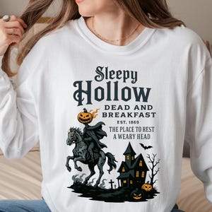 "Sleepy Hollow Halloween shirt featuring a vintage-style Headless Horseman on a spooky horse holding a jack-o'-lantern in front of a haunted house with glowing pumpkins and bats. This eerie Halloween graphic tee is printed on a moss green t-shirt and perfect for spooky season, trick-or-treat, horror movie lovers, gothic fall fashion, haunted house fans, or those searching for a unique October outfit. Ideal for fall events, pumpkin patches, or Halloween parties."