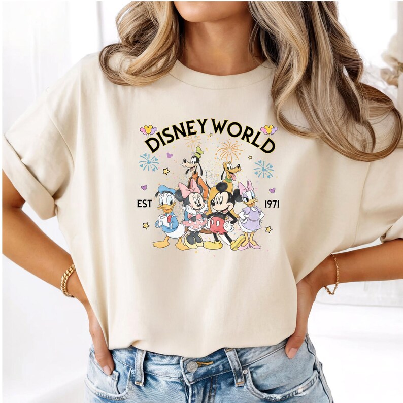 Puede incluir: Camiseta color crema con las palabras "Disney World" sobre un gr&aacute;fico de personajes de Disney, incluyendo Mickey Mouse, Minnie Mouse, Donald Duck y Goofy. Tambi&eacute;n incluye el texto "EST 1971".