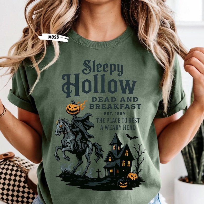 "Sleepy Hollow Halloween shirt featuring a vintage-style Headless Horseman on a spooky horse holding a jack-o'-lantern in front of a haunted house with glowing pumpkins and bats. This eerie Halloween graphic tee is printed on a moss green t-shirt and perfect for spooky season, trick-or-treat, horror movie lovers, gothic fall fashion, haunted house fans, or those searching for a unique October outfit. Ideal for fall events, pumpkin patches, or Halloween parties."