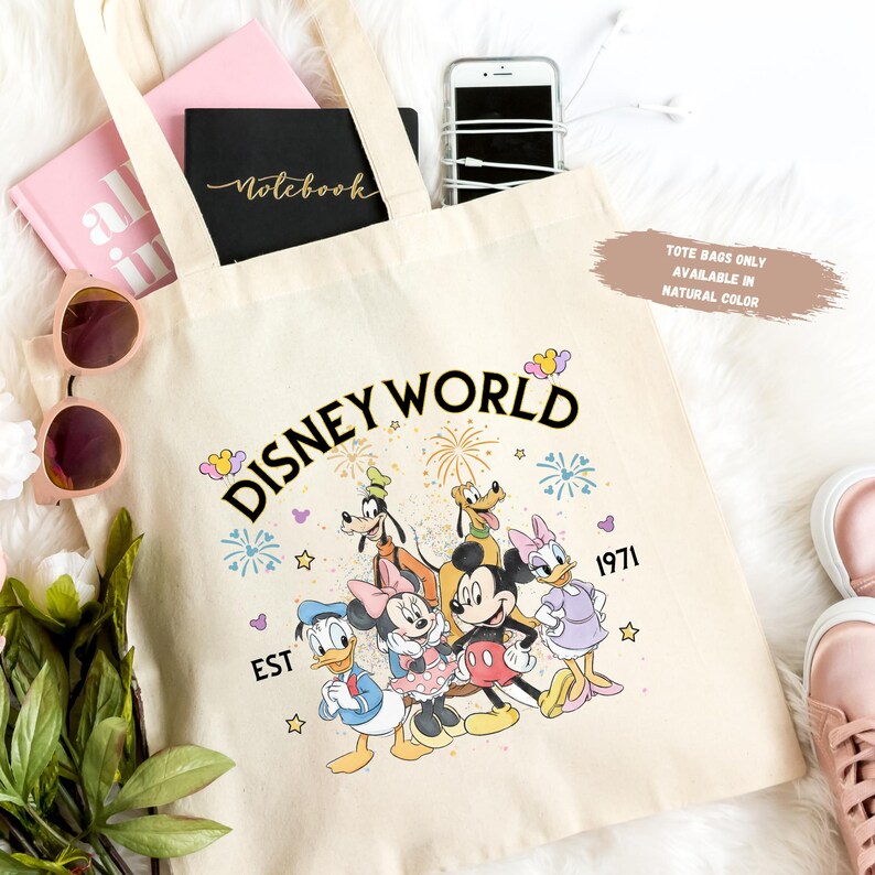 Puede incluir: Bolso tote de color crema con las palabras "Disney World" en negro, personajes de dibujos animados coloridos y el a&ntilde;o "1971". La bolsa contiene un cuaderno rosa, un cuaderno negro, un tel&eacute;fono y gafas de sol.