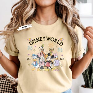 Puede incluir: Camiseta amarillo claro con la inscripci&oacute;n "Disney World" y un gr&aacute;fico de personajes de Disney: Mickey, Minnie, Donald, Daisy, Goofy y Pluto. El texto "EST 1971" est&aacute; debajo.