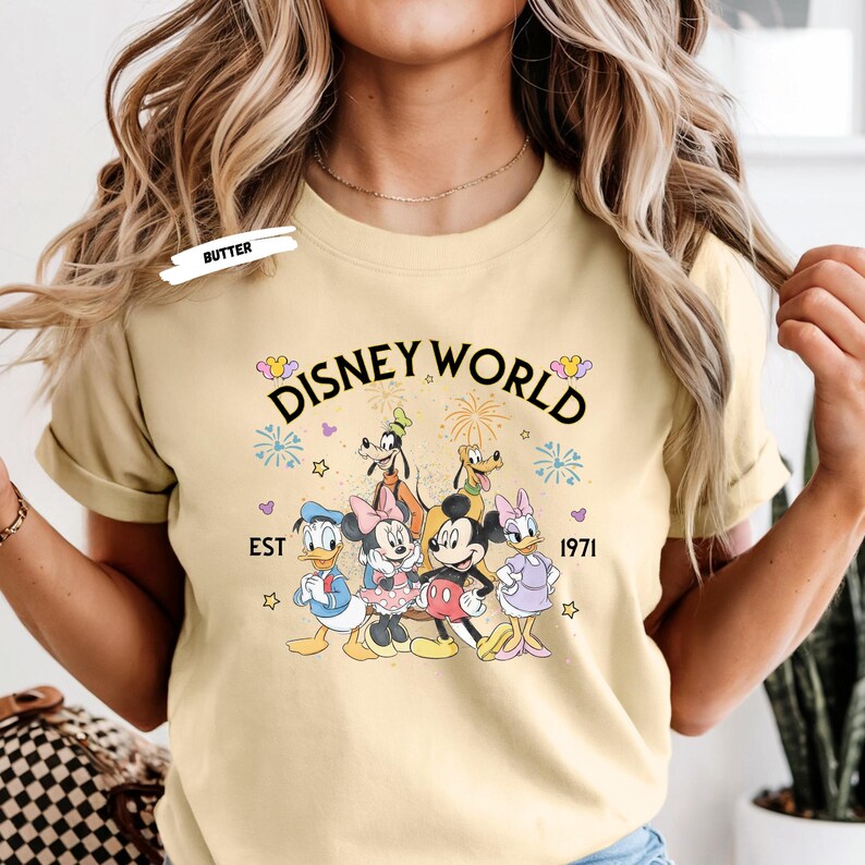 Puede incluir: Camiseta amarillo claro con la inscripci&oacute;n "Disney World" y un gr&aacute;fico de personajes de Disney: Mickey, Minnie, Donald, Daisy, Goofy y Pluto. El texto "EST 1971" est&aacute; debajo.
