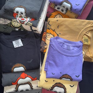 May include: A stack of folded t-shirts in various colors, including black, gray, purple, and yellow. Each shirt features a graphic of a horror movie character. Some shirts have bat and star designs.