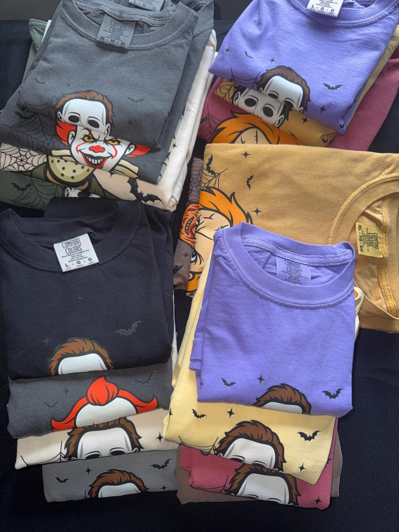 May include: A stack of folded t-shirts in various colors, including black, gray, purple, and yellow. Each shirt features a graphic of a horror movie character. Some shirts have bat and star designs.