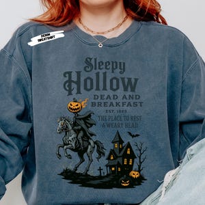"Sleepy Hollow Halloween shirt featuring a vintage-style Headless Horseman on a spooky horse holding a jack-o'-lantern in front of a haunted house with glowing pumpkins and bats. This eerie Halloween graphic tee is printed on a moss green t-shirt and perfect for spooky season, trick-or-treat, horror movie lovers, gothic fall fashion, haunted house fans, or those searching for a unique October outfit. Ideal for fall events, pumpkin patches, or Halloween parties."