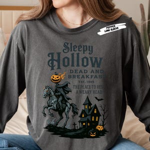 "Sleepy Hollow Halloween shirt featuring a vintage-style Headless Horseman on a spooky horse holding a jack-o'-lantern in front of a haunted house with glowing pumpkins and bats. This eerie Halloween graphic tee is printed on a moss green t-shirt and perfect for spooky season, trick-or-treat, horror movie lovers, gothic fall fashion, haunted house fans, or those searching for a unique October outfit. Ideal for fall events, pumpkin patches, or Halloween parties."