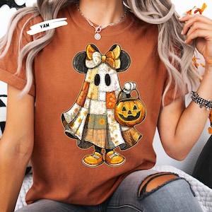 May include: Rust-colored t-shirt featuring a Halloween-themed graphic. The graphic depicts a ghost character with Mickey Mouse ears, a bow, and a patchwork design. The ghost holds a jack-o'-lantern. The word "YAM" is in the upper left corner.