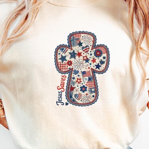 Puede incluir: Camiseta de color marfil con un diseño de cruz patriótica. La cruz está llena de estrellas, fuegos artificiales y banderas rojas, blancas y azules. Las palabras "Jesus Saves" están impresas verticalmente en el lado izquierdo de la cruz.
