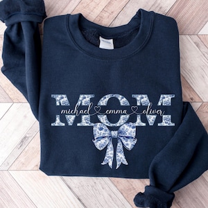 May include: Navy blue sweatshirt with the word "MOM" in a white and blue floral pattern. Below the word "MOM" are the names "michael", "emma", and "oliver". A blue and white floral bow is centered below the names.