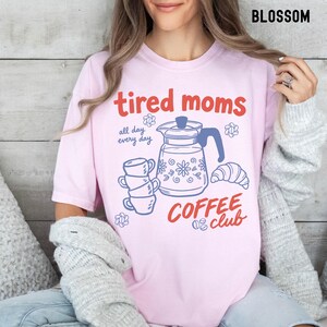 Vintage style graphic t-shirt featuring coffee pot, stacked cups, croissant, and playful tired moms coffee club text in soft pastel colors for moms. Printed on Comfort Colors t-shirt in Blossom.