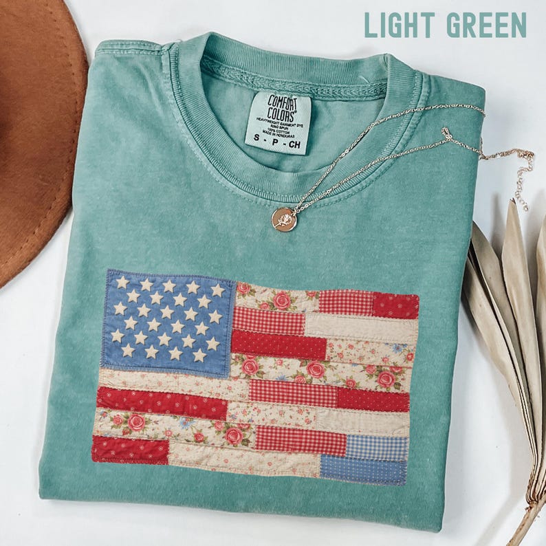 May include: Light green Comfort Colors t-shirt with a patchwork American flag design. The flag features blue stars, red and floral stripes. A silver necklace with a pendant rests on the shirt. The shirt is size S-P-CH.