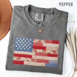 May include: A gray Comfort Colors t-shirt with a patchwork American flag design. The flag features blue stars, red and white stripes, and floral patterns. A silver necklace with a pendant is draped on the shirt.