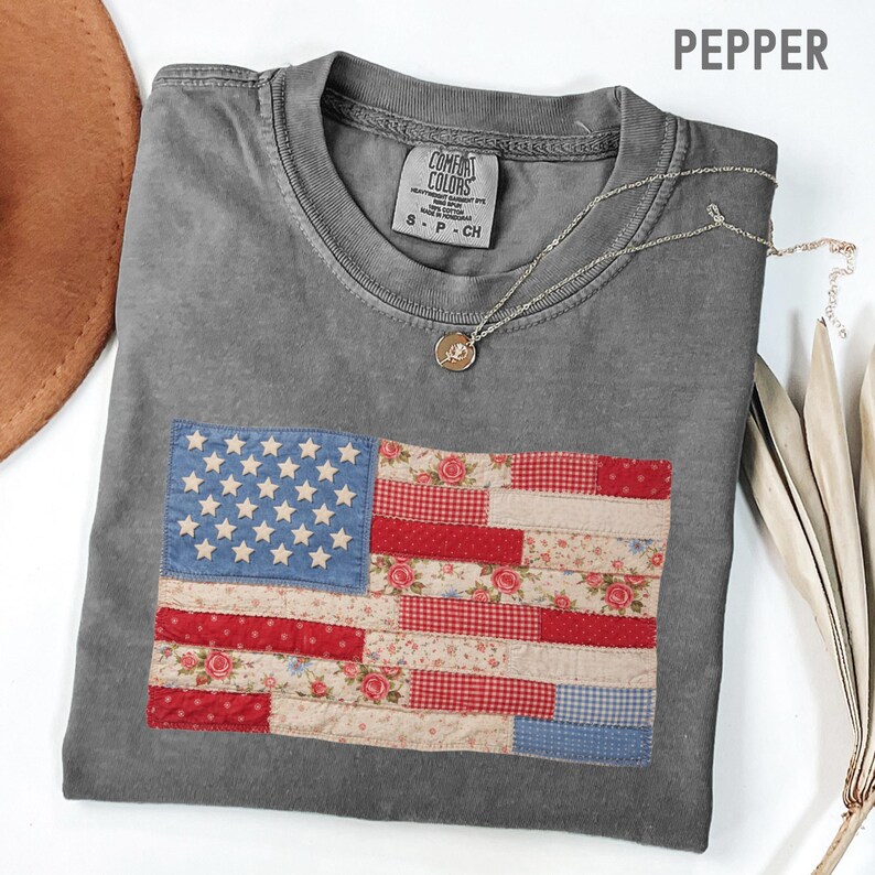 May include: A gray Comfort Colors t-shirt with a patchwork American flag design. The flag features blue stars, red and white stripes, and floral patterns. A silver necklace with a pendant is draped on the shirt.
