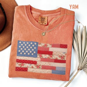 May include: Coral-colored Comfort Colors t-shirt featuring a patchwork American flag design. The flag incorporates blue, red, and floral patterned fabrics. A gold necklace with a pendant rests on the shirt. A brown hat and a dried palm leaf are also visible.