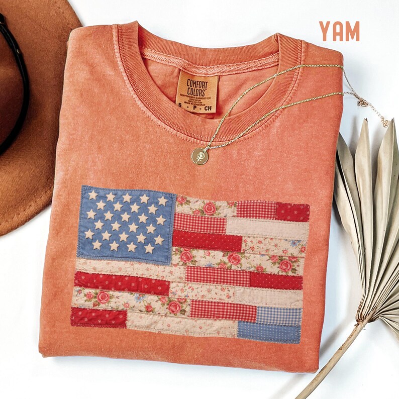 May include: Coral-colored Comfort Colors t-shirt featuring a patchwork American flag design. The flag incorporates blue, red, and floral patterned fabrics. A gold necklace with a pendant rests on the shirt. A brown hat and a dried palm leaf are also visible.