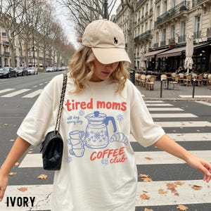 Vintage style graphic t-shirt featuring coffee pot, stacked cups, croissant, and playful tired moms coffee club text in soft pastel colors for moms. Printed on Comfort Colors t-shirt in Ivory.