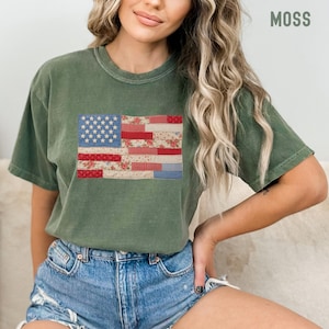 May include: Moss green t-shirt featuring a patchwork American flag design. The flag incorporates floral and gingham patterns in red, white, and blue. The word "MOSS" is printed in white on the upper right corner of the shirt.