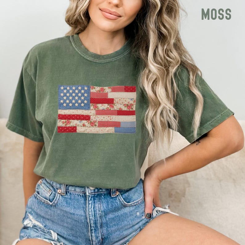 May include: Moss green t-shirt featuring a patchwork American flag design. The flag incorporates floral and gingham patterns in red, white, and blue. The word "MOSS" is printed in white on the upper right corner of the shirt.