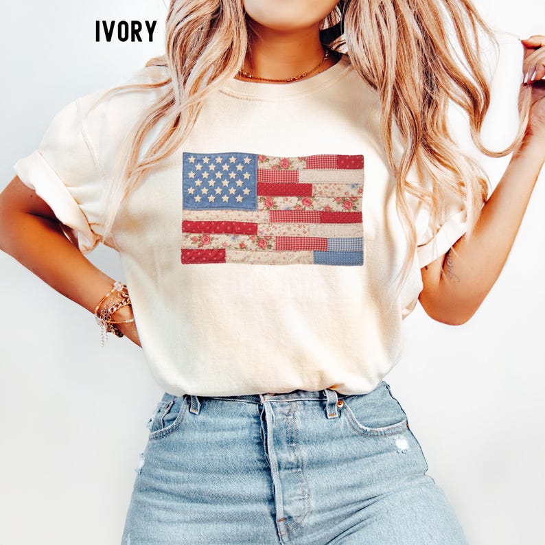 May include: Ivory-colored t-shirt featuring a patchwork American flag design. The flag incorporates blue stars, red and white stripes with floral and gingham patterns. The word "IVORY" is printed at the top of the shirt.