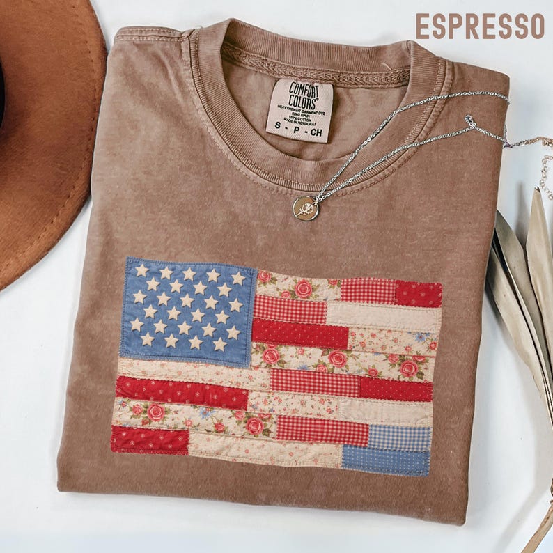May include: Espresso-colored t-shirt with a patchwork American flag design. The flag features blue stars, red and white stripes, and floral patterns. The shirt has a crew neck and short sleeves. A silver necklace with a pendant is draped on the shirt.