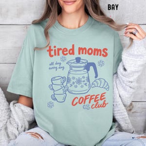 Vintage style graphic t-shirt featuring coffee pot, stacked cups, croissant, and playful tired moms coffee club text in soft pastel colors for moms. Printed on Comfort Colors t-shirt in Bay.