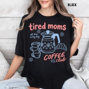 Vintage style graphic t-shirt featuring coffee pot, stacked cups, croissant, and playful tired moms coffee club text in soft pastel colors for moms. Printed on Comfort Colors t-shirt in Black.