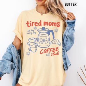 Vintage style graphic t-shirt featuring coffee pot, stacked cups, croissant, and playful tired moms coffee club text in soft pastel colors for moms. Printed on Comfort Colors t-shirt in Butter.