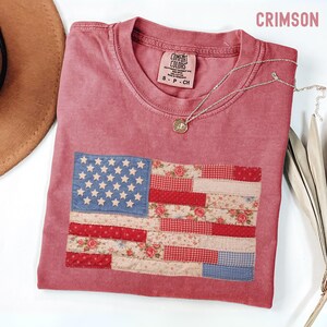 May include: Crimson-colored t-shirt with a patchwork American flag design. The flag features a blue field with white stars and red and floral-patterned stripes. A silver necklace with a pendant is draped on the shirt.
